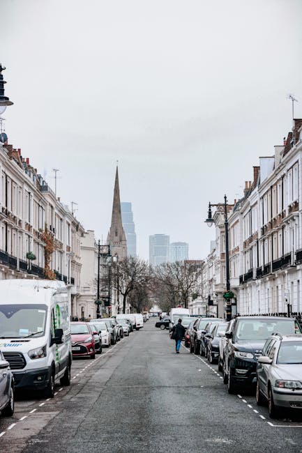 A photograph of a residential street in Shepherds Bush W12, showing rows of white and cream-colored terraced houses with black wrought-iron balconies and window railings. Parked cars line both sides of the asphalt road, including a white van and various sedans. At the street's end, a large church steeple rises above the tree canopy, with modern high-rise buildings visible in the background under a cloudy sky. The scene is well-lit with natural daylight, and the overall atmosphere is calm and orderly, reflecting typical urban residential architecture. This image exemplifies the clean and maintained appearance of urban streets, aligning with professional cleaning company's focus on surface cleaning and maintenance for homes in the Shepherds Bush area.