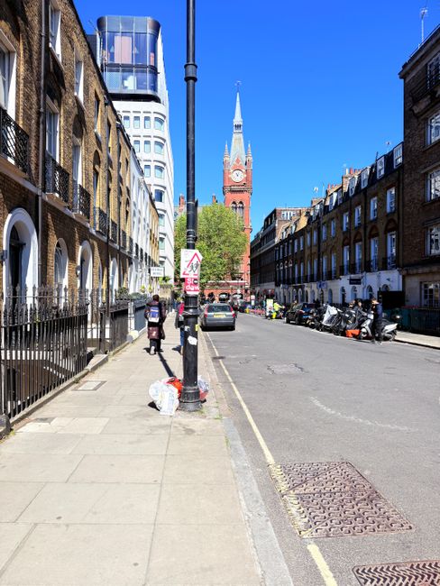 A sunny street scene in Shepherds Bush, featuring a row of terraced brick residential buildings on the right side and a modern glass skyscraper on the left. The foreground shows a clean pavement with a few pedestrians, some carrying bags, walking along the sidewalk. Parked motorcycles and cars line the curb, with a traffic sign and a lamppost visible in the middle ground. In the background, the red-brick clock tower of St. Mary’s Church stands prominently against a bright blue sky. The scene reflects urban cleanliness and maintenance typical of residential and commercial areas in Shepherds Bush as promoted by [COMPANY_NAME] for surface cleaning and hygienic upkeep.