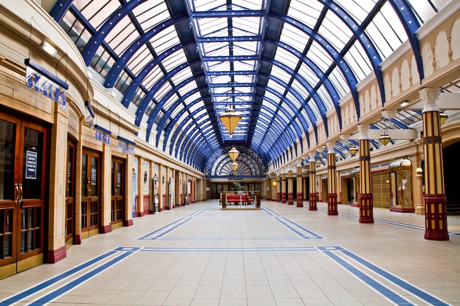 Interior view of a spacious shopping mall corridor featuring a high, arched glass ceiling with blue metal framework, allowing natural light to illuminate the area. The polished tiled floor is predominantly beige with blue decorative lines, creating a clean and bright appearance. On the left side, retail store entrances with wooden and glass doors are visible, some with signs and notices. Tall, white columns with gold and red accents support the ceiling, adding an elegant architectural detail. The corridor appears well-maintained and free of dust or debris, exemplifying thorough cleaning and sanitisation that [COMPANY_NAME] provides for commercial spaces like Shepherds Bush shopping centres, contributing to hygienic, inviting environments near Westfield Shepherds Bush.