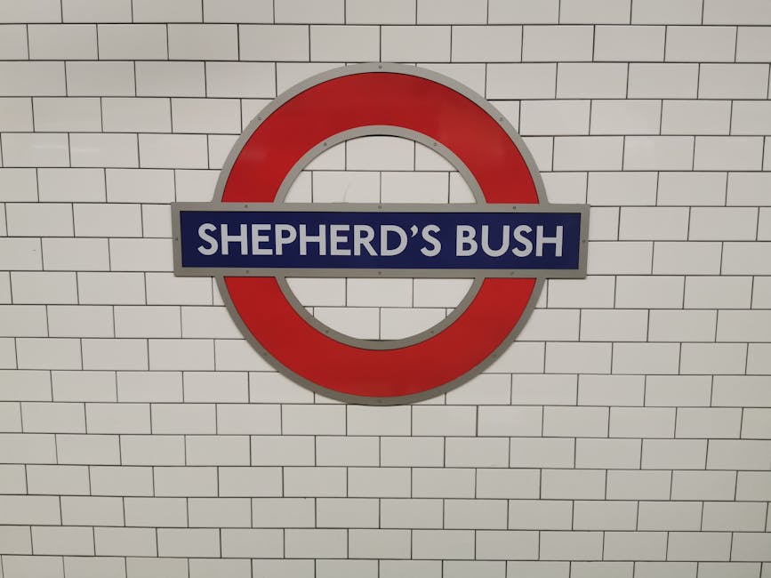 Close-up of a Shepherd's Bush London Underground station sign mounted on a white tiled wall, featuring a red circular border with a blue band across the center displaying the station name in white text. The tiles are uniformly arranged, clean, and glossy, reflecting well-maintained subway lighting. The sign's surface appears smooth and free of fingerprints or dirt, highlighting the precision of surface cleaning and sanitisation typical of professional cleaning services, such as those provided by [COMPANY_NAME], at the Uxbridge Road end of tenancy cleaning in Shepherds Bush. The background shows a well-lit, tidy subway wall surface, emphasizing hygiene and cleanliness in public transportation environments.
