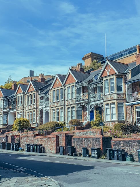A row of Victorian-style terraced houses on Uxbridge Road in Shepherds Bush, featuring ornate white ironwork balconies and bay windows. The brick facades are clean and well-maintained, with some houses having small front gardens bordered by low brick walls. The street in front is lined with black wheelie bins and a paved sidewalk, with clear blue skies overhead. The scene reflects a tidy, residential area, emphasizing the importance of surface cleaning and maintenance, as highlighted by [COMPANY_NAME] for end of tenancy cleaning in Shepherds Bush at https://carpetcleaningshepherdsbush.com.